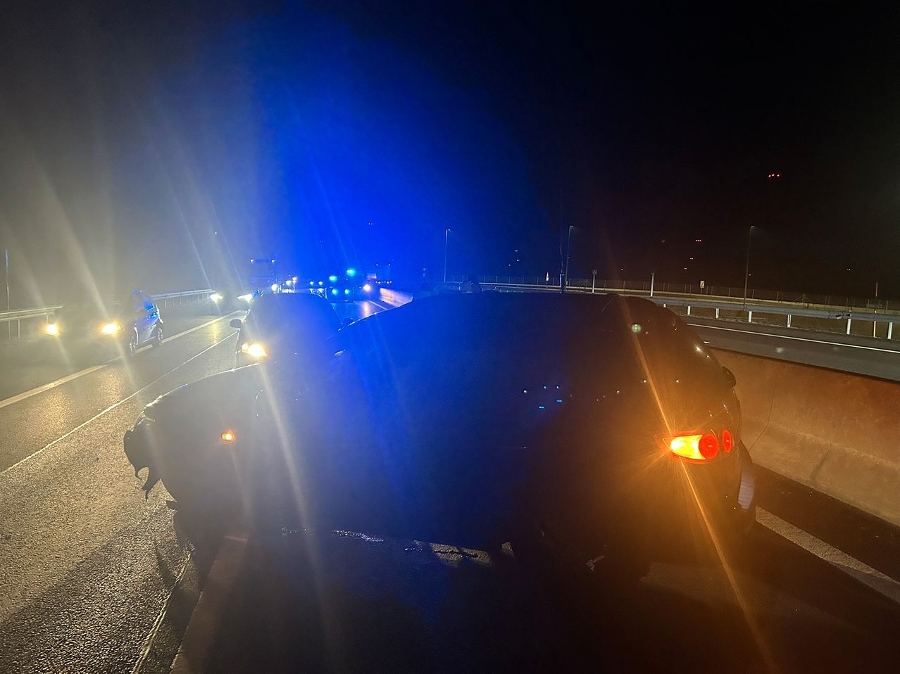 Rettungsfahrzeuge stehen nachts auf einer Brücke. Die Rückseite eines Autos ist von blauen Lichtern beleuchtet.