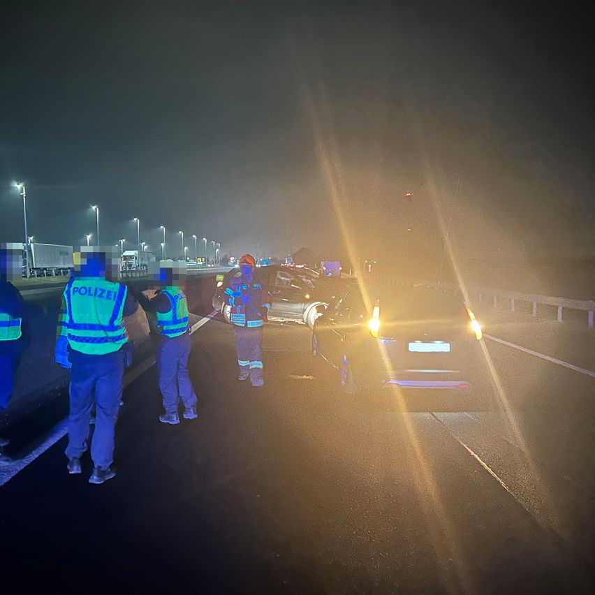 Polizisten in Warnwesten untersuchen einen Autounfall auf einer Autobahn bei Nacht. Das Auto steht mit eingeschalteten Warnblinkleuchten am Straßenrand.