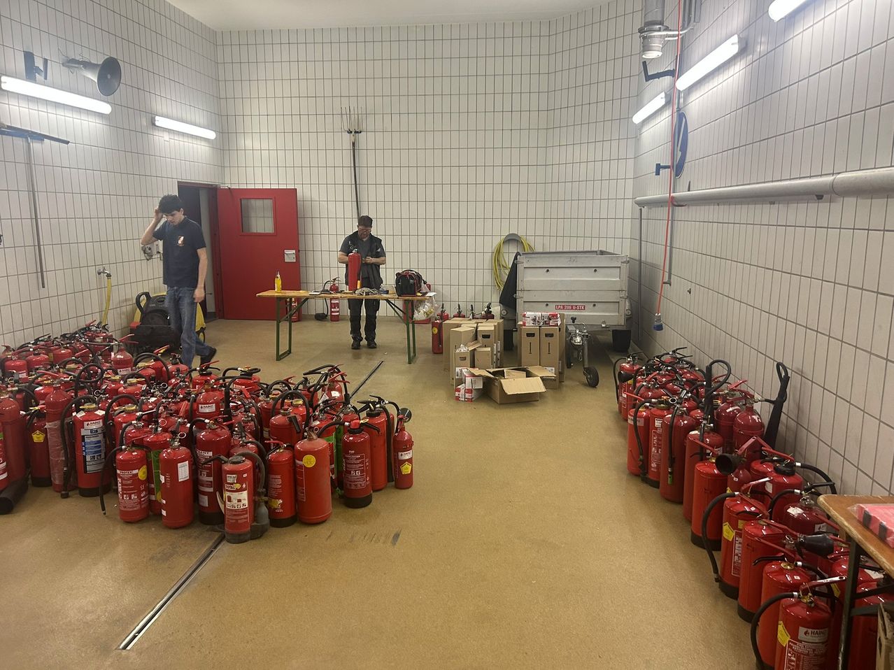 Zwei Personen arbeiten in einem Raum mit gefliesten Wänden an einem Tisch mit Feuerlöschern und anderem Equipment. Mehrere rote Feuerlöscher sind auf beiden Seiten aufgestellt.
