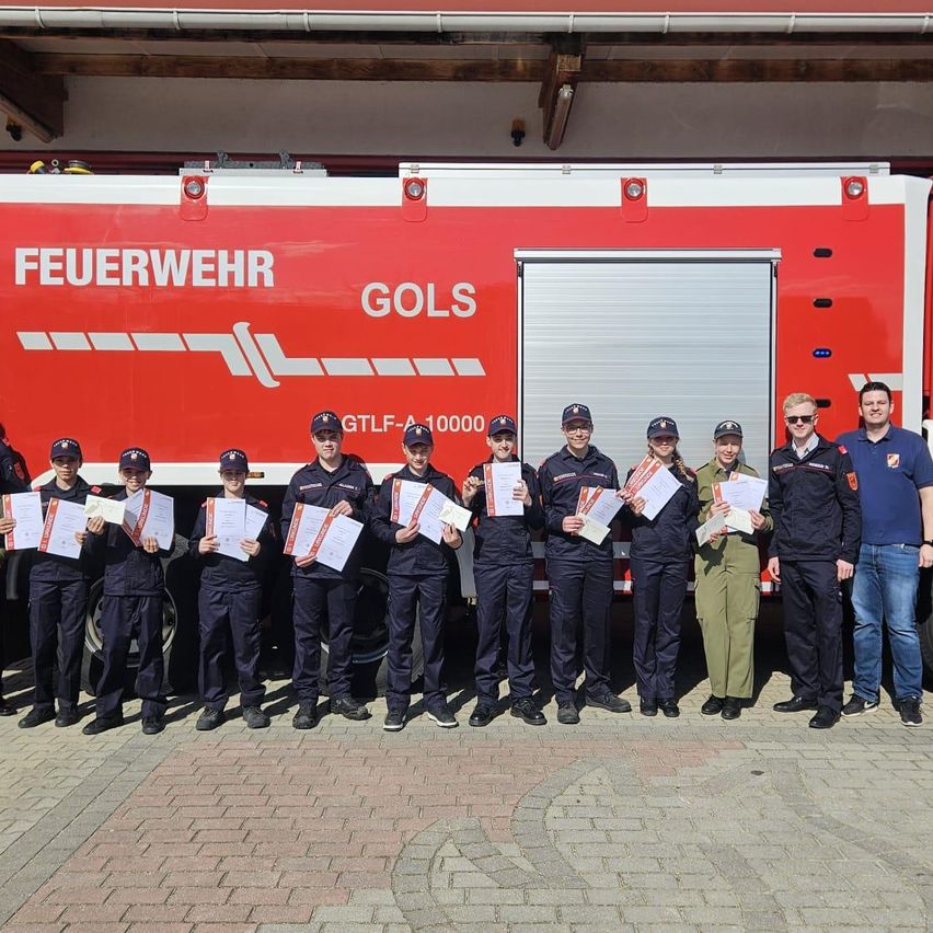 Eine Gruppe Feuerwehrleute steht vor einem roten Feuerwehrauto. Sie tragen alle Uniformen und halten Zertifikate. Sie posieren für ein Foto.