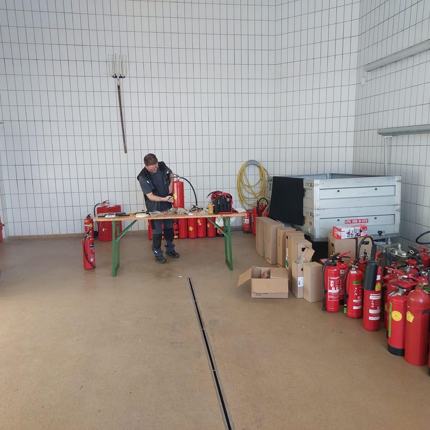 Ein Mann in Sicherheitsausrüstung arbeitet an einem Tisch mit Feuerlöschern in einem Lagerraum. Kisten und weitere Feuerlöscher sind auf dem Boden verstreut.