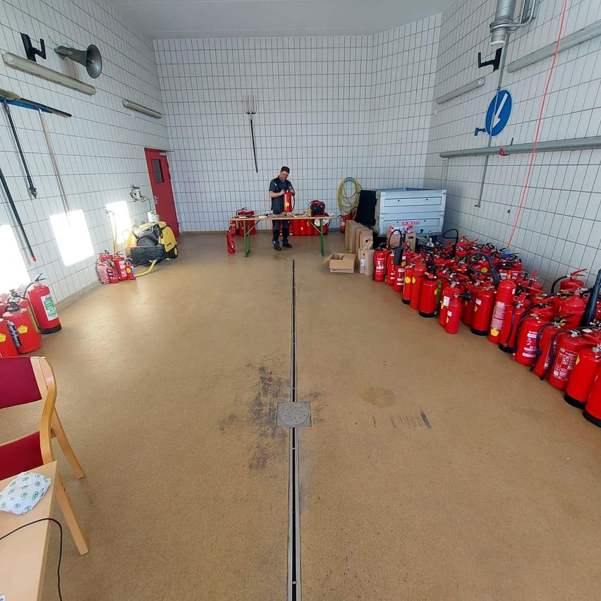 Ein Mann steht in einem Raum mit weißen Fliesenwänden und hält einen Feuerlöscher. Rote Feuerlöscher säumen die Wand, und ein Tisch mit verschiedenen Gegenständen befindet sich in der Mitte.