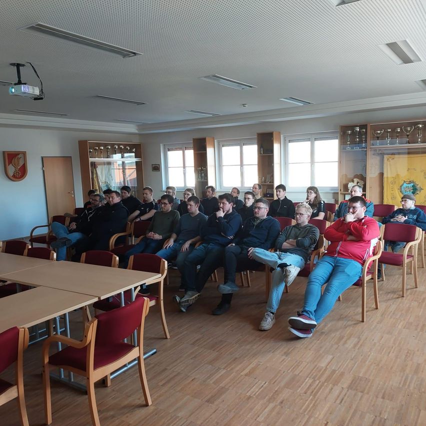 Ein Raum mit Holzboden und Stühlen, einer Projektor und Deckenleuchten. Mehrere Personen sitzen in Reihen, einige mit Brillen und Turnschuhen. Banner und Trophäen schmücken den Raum.