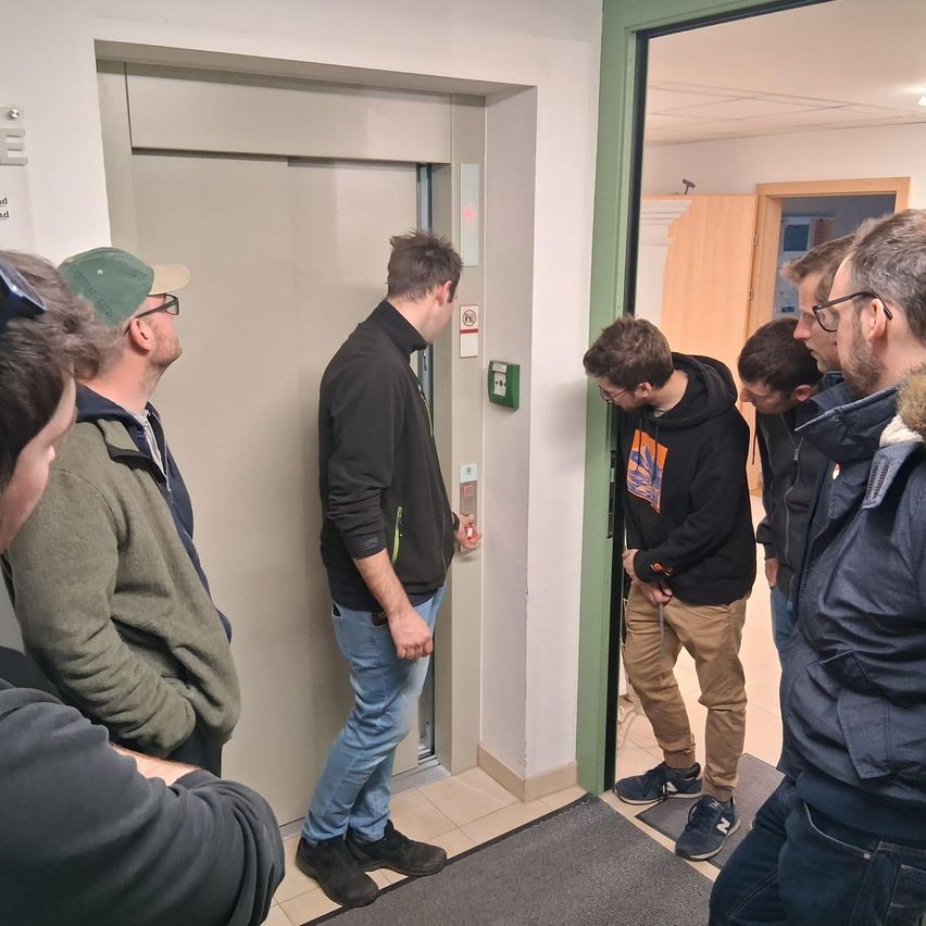 Eine Gruppe von Männern steht in einem Flur. Ein Mann drückt einen Aufzugsknopf. Ein anderer Mann verlässt den Aufzug.