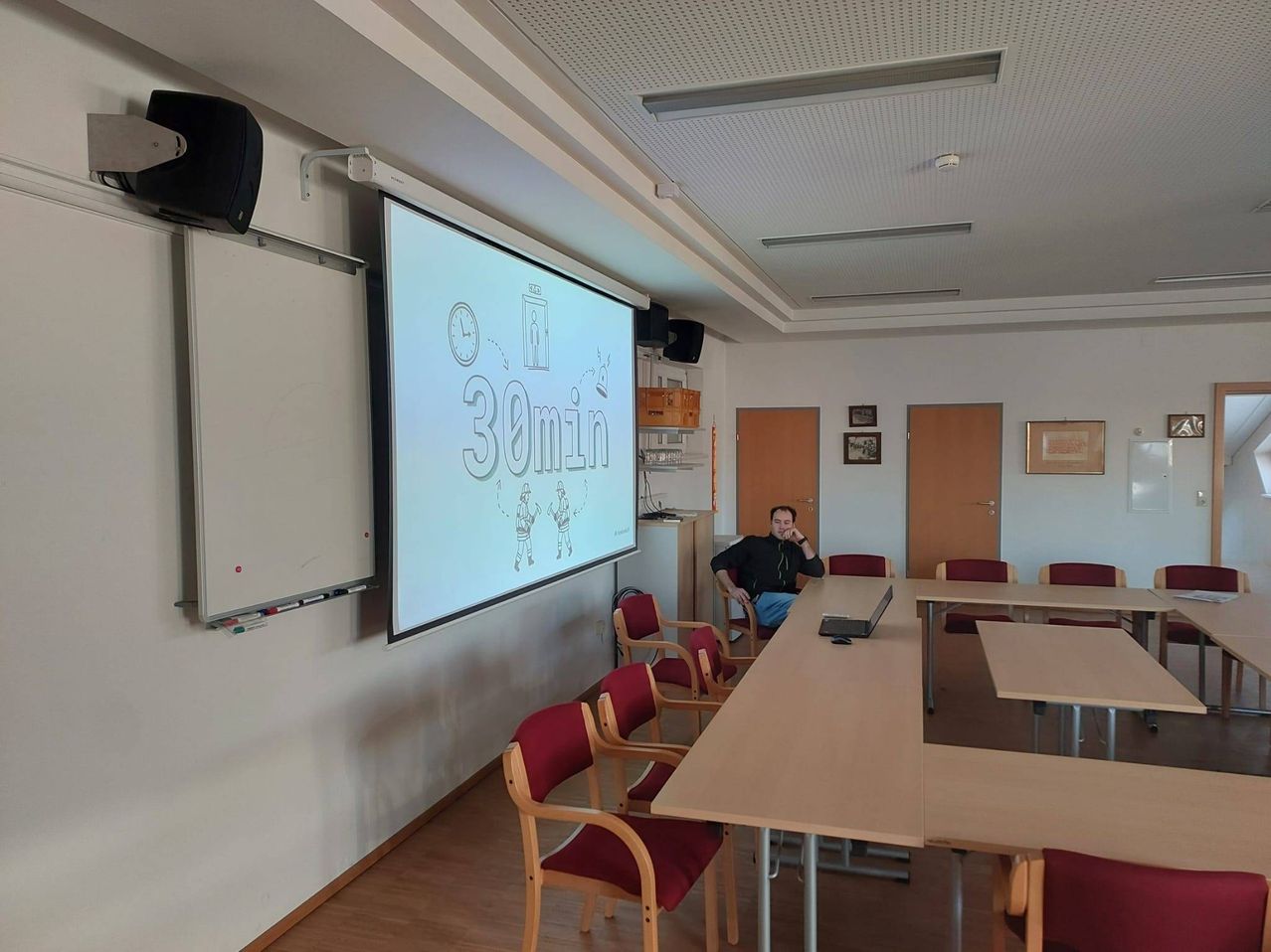 Ein Mann sitzt an einem Tisch in einem Konferenzraum und schaut auf einen Bildschirm mit 30min. Der Raum hat weiße Wände, Holzböden und eine Tafel.