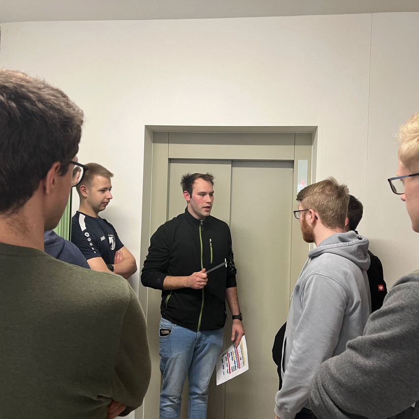 Ein Mann steht vor einer Gruppe von Männern und hält ein Papier in der Hand. Er erklärt ihnen etwas. Sie stehen vor einer Tür.