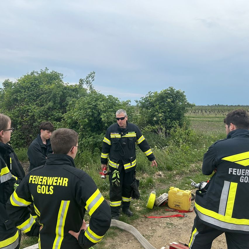 Eine Gruppe von Feuerwehrleuten versammelt sich auf einem offenen Feld. Einer von ihnen trägt eine Sonnenbrille und ein schwarzes Uniform. Sie stehen um einen Feuerlöscher und einen gelben Kanister herum.