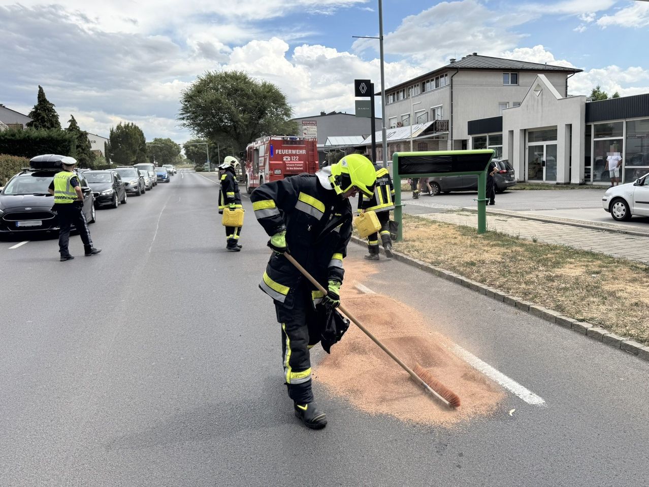 Feuerwehrleute in Schutzkleidung reinigen mit einem Besen eine Straßenverschmutzung, während ein Feuerwehrwagen in der Nähe geparkt ist.