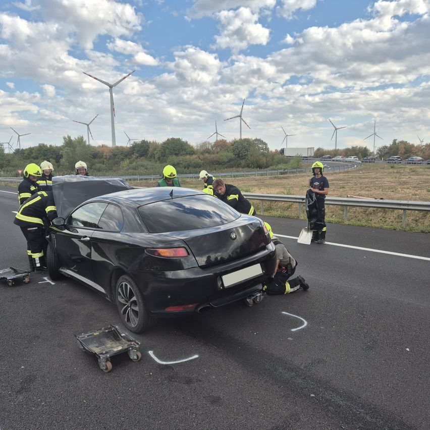 Rettungskräfte in gelben Helmen arbeiten an einem beschädigten schwarzen Auto am Straßenrand, mit Windturbinen im Hintergrund.