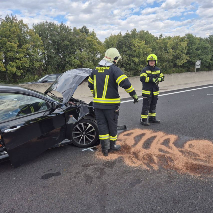 Zwei Feuerwehrleute in voller Ausrüstung stehen auf einer Autobahn, einer in der Nähe eines beschädigten schwarzen Autos mit offenem Motorhaube und der andere in der Nähe einer Sandverschüttung.