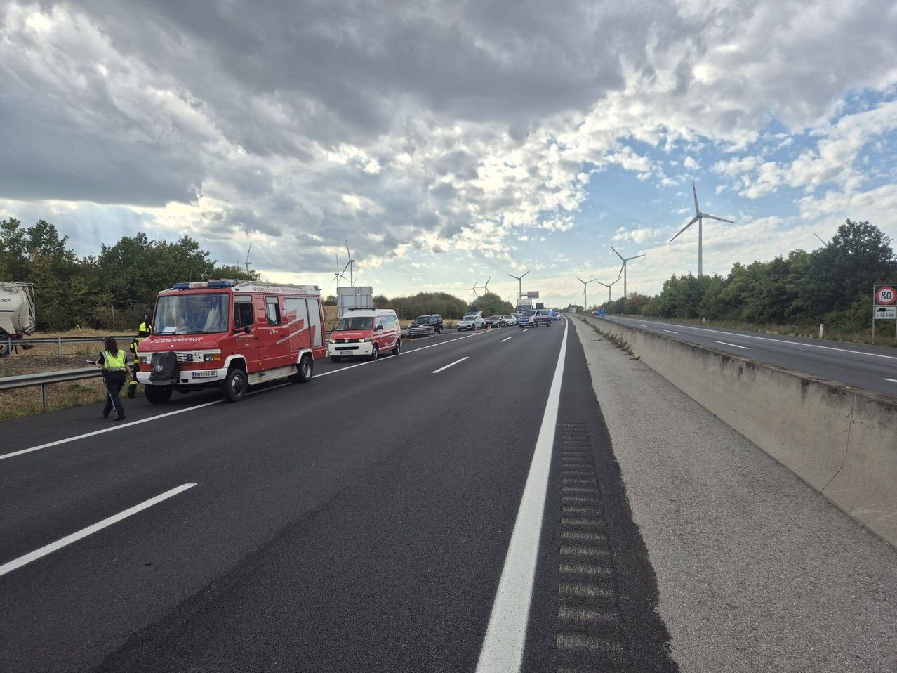 Einsatzfahrzeuge stehen am Straßenrand einer Autobahn, während der Verkehr in die entgegengesetzte Richtung fährt. Im Hintergrund sind mehrere Windräder zu sehen.