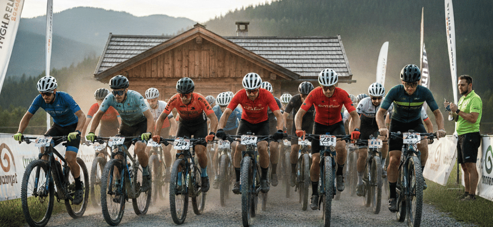 Eine Gruppe von Radfahrern mit Helmen und Radsportbekleidung nimmt an einem Rennen auf einer Schotterstraße teil. Hinter ihnen steht ein Holzgebäude mit Schornstein, umgeben von Fahnen und einem Rasenbereich. Berge sind im Hintergrund zu sehen.