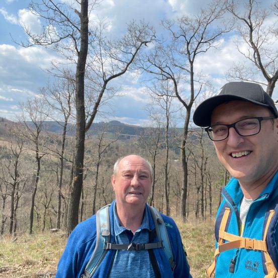 Zwei Männer stehen in einem Wald. Einer trägt eine blaue Jacke und einen Rucksack, der andere eine schwarze Mütze und eine Brille.
