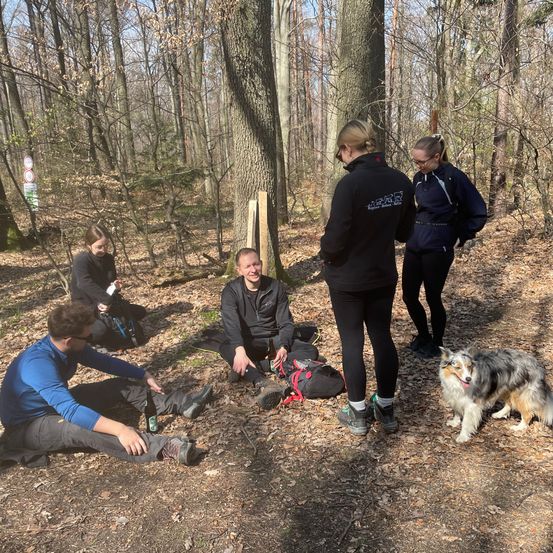 Vier Personen sitzen auf dem Boden in einem Wald. Eine Person steht vor ihnen. Ein Hund steht vor ihnen.