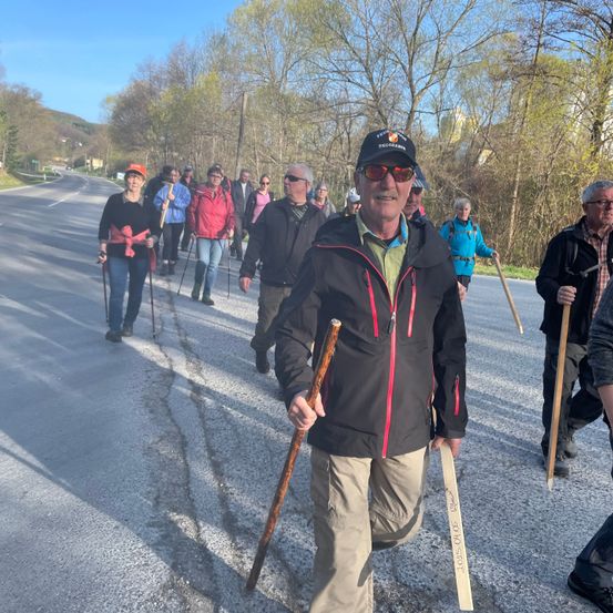 Eine Gruppe von Menschen geht auf einem Schotterweg und hält Stöcke. Ein Mann in einer schwarzen Jacke mit roten Reißverschlüssen geht vorne. Die Bäume auf beiden Seiten der Straße sind grün. Die Schatten der Menschen fallen auf die Straße. Der Himmel ist blau.