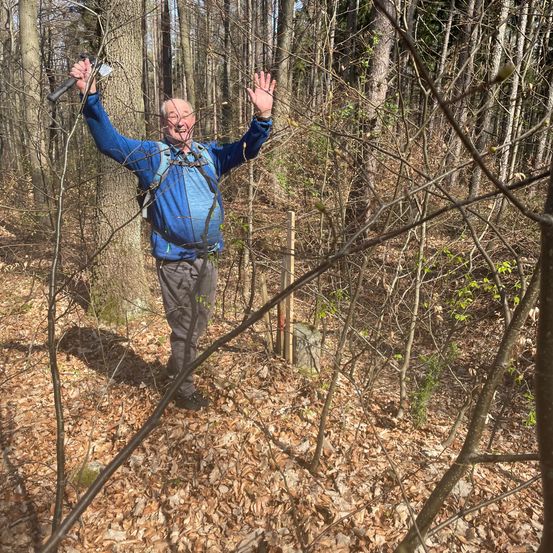 Ein älterer Mann in einem blauen Jacket und grauen Hosen steht im Wald, mit erhobenen Händen und einem Holzstock in der linken Hand.