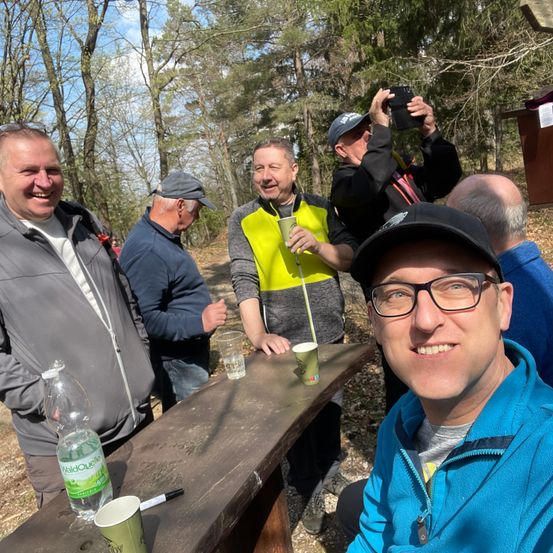 Gruppe lächelnder Männer in einer Outdoor-Umgebung, einer nimmt ein Foto auf, die anderen sitzen um einen Holztisch mit Tassen und Flaschen.