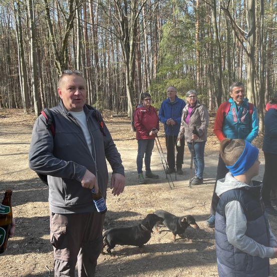 Eine Gruppe von Menschen und Hunden steht in einem Wald. Ein Mann in einer grauen Jacke hält eine Flasche Bier, und zwei kleine schwarze Hunde gehen herum.