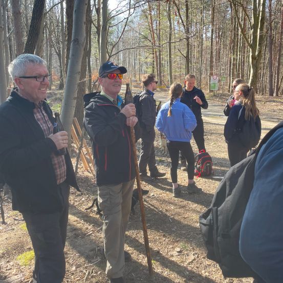 Eine Gruppe von Menschen steht in einem Wald, ein Mann hält einen Wanderstock, ein anderer Mann hat einen Rucksack, eine Frau hat geflochtene Haare und ein Mann redet mit einem anderen Mann.