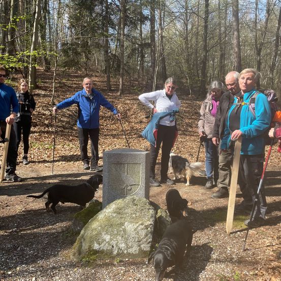 Eine Gruppe von Wanderern steht in einem Waldgebiet, mit einem Steinmarker in der Mitte, und einige Hunde begleiten sie.
