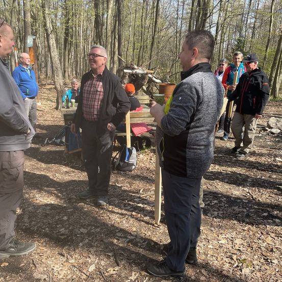 Eine Gruppe von Menschen steht in einem Waldgebiet und einige halten Stöcke. Ein Mann mit Brille und kariertem Hemd steht vor einem anderen Mann in einer grauen Jacke. Sie scheinen zu sprechen. Eine Holzbank ist hinter ihnen aufgestellt. Einige Leute sitzen auf der Bank.