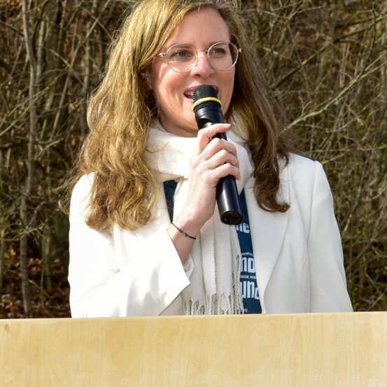 Eine Frau mit blonden Haaren, die eine Brille, einen weißen Mantel und einen weißen Schal trägt, steht hinter einem hölzernen Podium und spricht in ein Mikrofon.