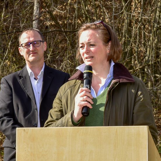 Eine Frau in einem grünen Shirt und einer Jacke spricht in ein Mikrofon an einem Rednerpult. Ein Mann in einem Anzug steht hinter ihr.