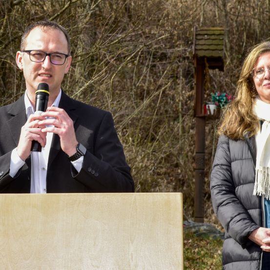 Ein Mann in einem Anzug hält ein Mikrofon und steht an einem Rednerpult. Eine Frau steht neben ihm und lächelt. Dahinter steht ein hölzernes Vogelhaus mit Blumen auf einem Pfahl.