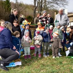 Eine Gruppe von Kindern und Erwachsenen in einem Park, einige stehen und andere sitzen. Eine Frau kniet und hält ein blaues Papier. Die Kinder tragen Mützen und Stiefel. Der Hintergrund zeigt einen Holzzaun und Bäume.