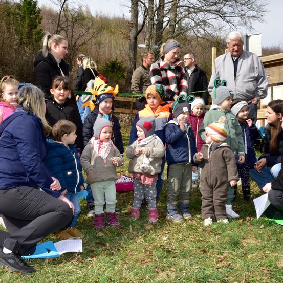 Eine Gruppe von Kindern und Erwachsenen in einem Park, einige stehen und andere sitzen. Eine Frau kniet und hält ein blaues Papier. Die Kinder tragen Mützen und Stiefel. Der Hintergrund zeigt einen Holzzaun und Bäume.
