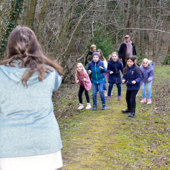 Eine Gruppe junger Mädchen und zwei Erwachsene gehen auf einem Waldweg. Die Mädchen sind in Winterkleidung gekleidet, einige in Stiefeln, andere in Turnschuhen. Die Erwachsenen sind hinter den Kindern, einer von ihnen trägt einen Hut.