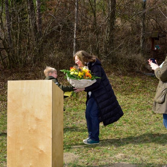 Eine Frau mit einer Blumenstrauß und lächelnd steht neben einem Holzpodium. Ein Kind mit einem Blumenstrauß steht hinter dem Podium. Ein Mann steht neben ihnen und hält ein Handy.