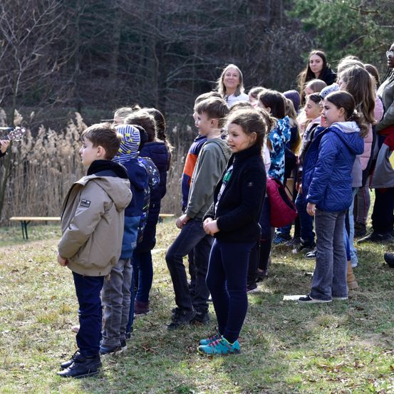 Eine Gruppe von Schülern steht draußen und hört einer Frau zu, die spricht. Einige Schüler schauen zur Frau, andere zum Boden.