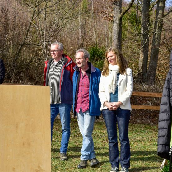 Drei Erwachsene stehen gemeinsam vor einem Holzbrett in einem Park. Hinter ihnen sind Bäume und ein Holzzaun.