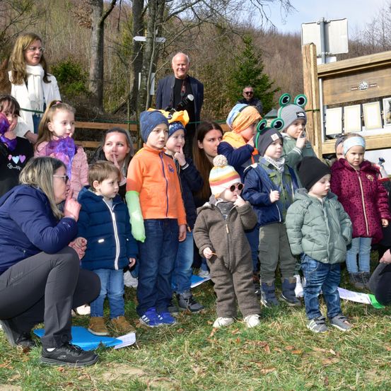 Eine Gruppe von Kindern und Erwachsenen steht in einem Außenbereich. Sie scheinen für ein Foto zu posieren. Sie tragen Winterkleidung. Die Kinder tragen Mützen und Schuhe. Eine Frau vorne kniet und hält ein Papier. Hinter ihnen steht ein Holzzaun.