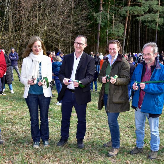 Vier Menschen stehen in einem Wald, lächeln und halten grüne Bänder. Die Frau auf der linken Seite trägt einen weißen Mantel, während der Mann neben ihr einen schwarzen Anzug trägt. Die Frau auf der rechten Seite ist in einem grünen Mantel und hält ein grünes Band. Dahinter stehen viele Menschen, und der Wald ist mit Bäumen gefüllt.
