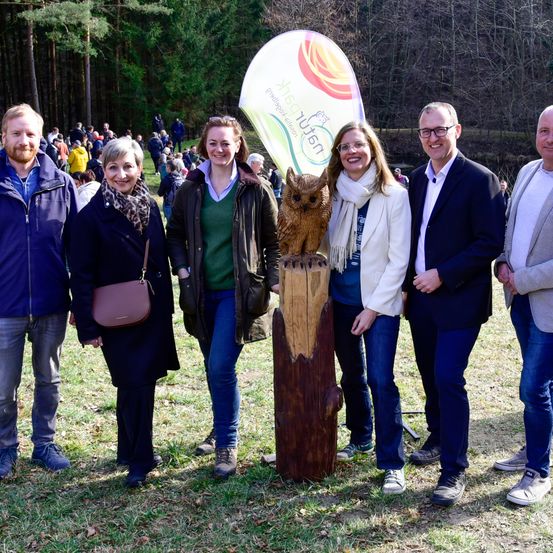 Eine Gruppe von Menschen steht vor einem hölzernen Pfosten mit einem geschnitzten Uhu. Sie alle lächeln und posieren für ein Foto.