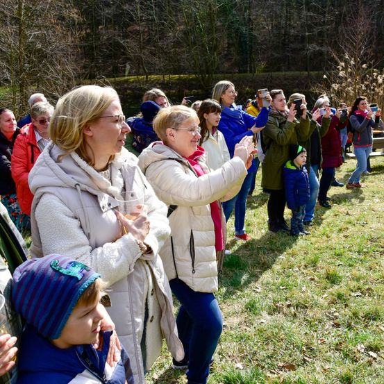 Eine Gruppe von Menschen steht auf einem Feld mit Bäumen im Hintergrund. Sie machen Fotos und Videos mit ihren Handys.