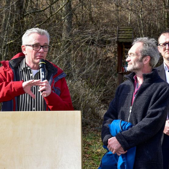 Drei Männer in einer Waldlichtung. Der Mann in der roten Jacke spricht in ein Mikrofon.