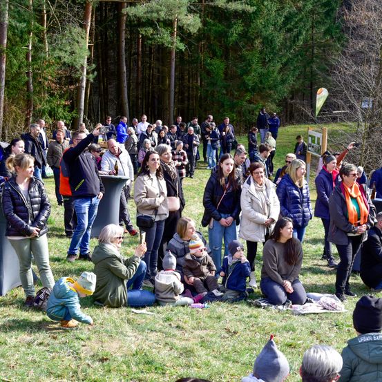 Eine große Gruppe von Menschen versammelt sich in einem Park. Einige stehen, andere sitzen und wieder andere sind auf dem Boden. Es gibt Menschen jeden Alters, einschließlich Kinder. Der Park ist von Bäumen umgeben.