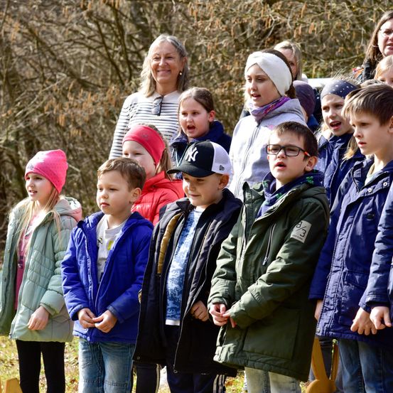 Eine Gruppe von Kindern in Winterkleidung, die in einer Reihe stehen und nach oben schauen. Eine Frau steht hinter ihnen und lächelt.
