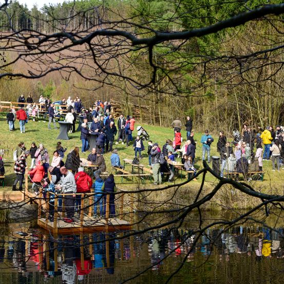 Eine große Gruppe von Menschen versammelt sich auf einem Rasenfeld, einige sitzen an Picknicktischen, andere stehen und unterhalten sich. Das Gebiet ist von Bäumen umgeben und es gibt eine Holzbrücke über einem Gewässer.