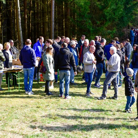 Eine Gruppe von Menschen steht auf einer Grasfläche, wahrscheinlich bei einer Veranstaltung. Einige tragen Jacken, Brillen und Sneakers. Ein Tisch mit Essen und Getränken steht im Vordergrund. Im Hintergrund sind mehrere Menschen zu sehen, einige tragen Hüte.