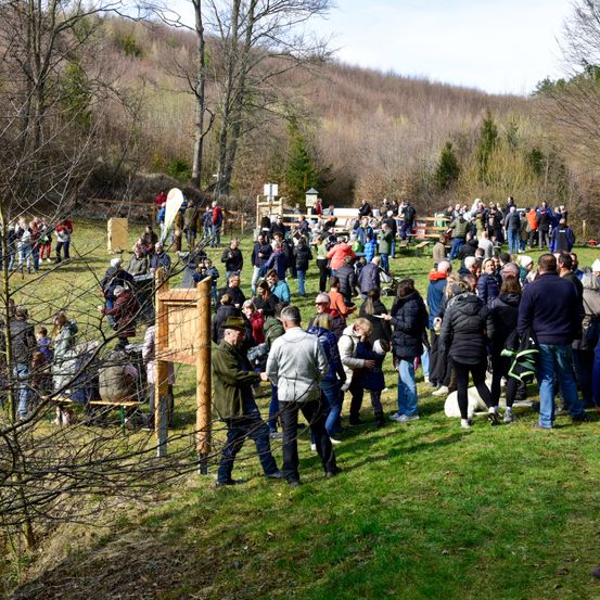 Eine große Gruppe von Menschen versammelt sich auf einer grasbewachsenen Wiese, einige gehen, andere stehen und unterhalten sich. Im Vordergrund befindet sich eine Holzstruktur, auf die ein Mann zugeht. Im Hintergrund zeigt sich eine hügelige Landschaft mit Bäumen und einem klaren Himmel.
