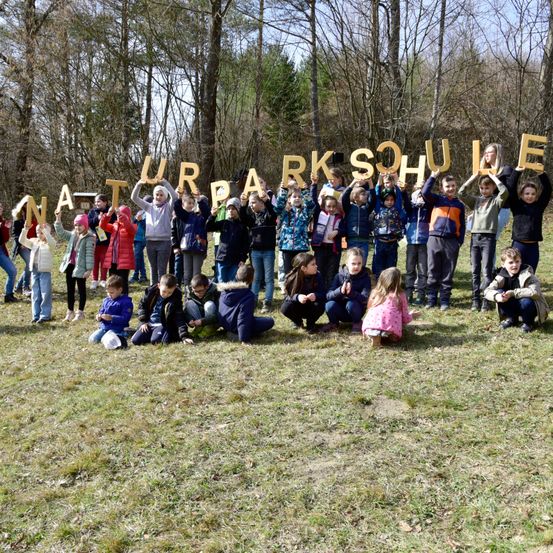 Gruppe von Schülern posiert für ein Foto vor einem Wald mit Buchstaben, die "NATURPARKSCHULE" buchstabieren.