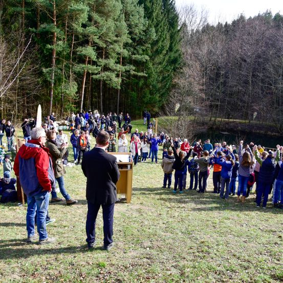 Eine Gruppe von Menschen versammelt sich in einer Waldlichtung, einige stehend und andere sitzend. Ein Mann in einem schwarzen Anzug steht an einem Podium und wendet sich an die Menge. Ein Fluss fließt hinter den Menschen, und sie scheinen an einer Zeremonie oder Veranstaltung teilzunehmen.