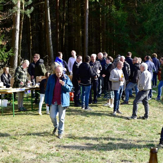 Eine Gruppe von Menschen versammelt sich in einem Wald, einige stehen und andere gehen. Ein Mann in einer blauen Jacke geht nach vorne. Im Vordergrund steht ein Tisch mit Essen, um den sich mehrere Menschen versammelt haben.