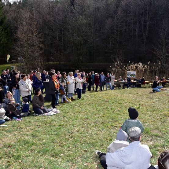 Eine Gruppe von Menschen versammelt sich in einer Freiluftveranstaltung, möglicherweise für ein Event, mit einigen, die auf dem Gras sitzen, und anderen, die stehen. Hinter ihnen sind Tische und Bänke aufgestellt, an denen einige Personen sitzen. Im Hintergrund befinden sich Bäume und ein See.