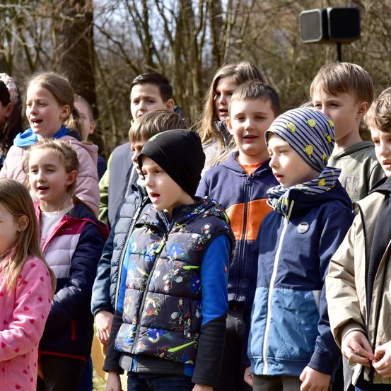 Eine Gruppe von Kindern, von denen einige Jacken tragen, steht in einem Waldgebiet und scheint zu singen.