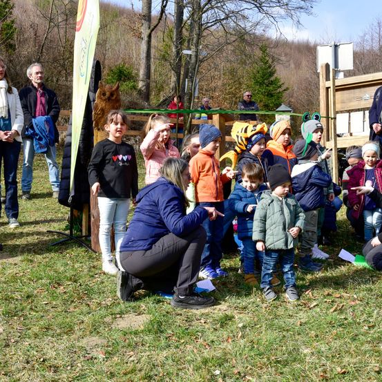 Eine Gruppe von Kindern und Erwachsenen steht in einem Außenbereich, wahrscheinlich in einem Park. Eine Frau kniet und zeigt auf etwas, während ein Kind es betrachtet. Eine Holzstruktur steht hinter ihnen, und ein Banner steht auf der linken Seite. In der Ferne gibt es Bäume und einige Menschen, die stehen.